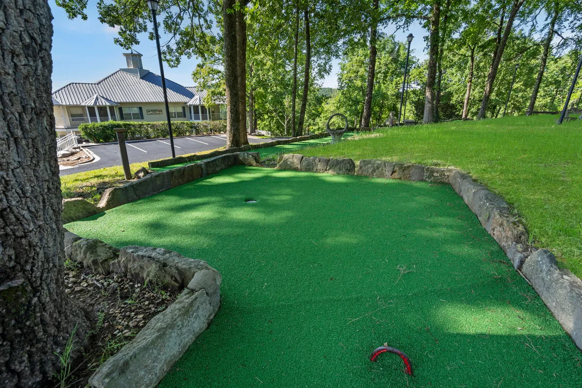 A playful mini-golf area with green synthetic turf and stone borders, nestled among trees. The inviting atmosphere suggests a setting for friendly competition and family togetherness outdoors.
