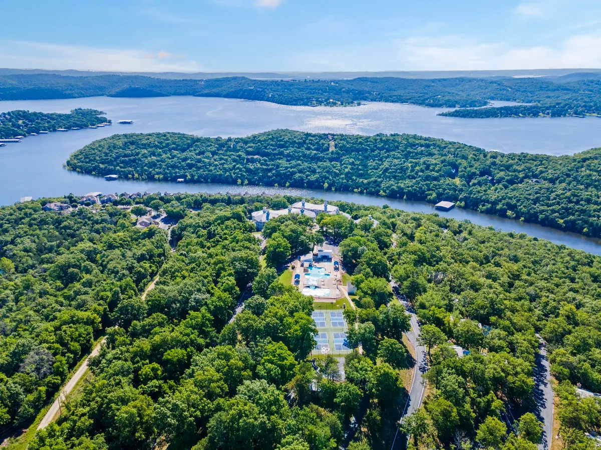 This wide view captures lush greenery, a shimmering lake, visible property amenities such as a swimming pool, and tennis courts. It’s a lively yet peaceful scene with space for water sports, tennis matches, and relaxation by the pool. The sun is setting, casting a golden glow over the whole area.