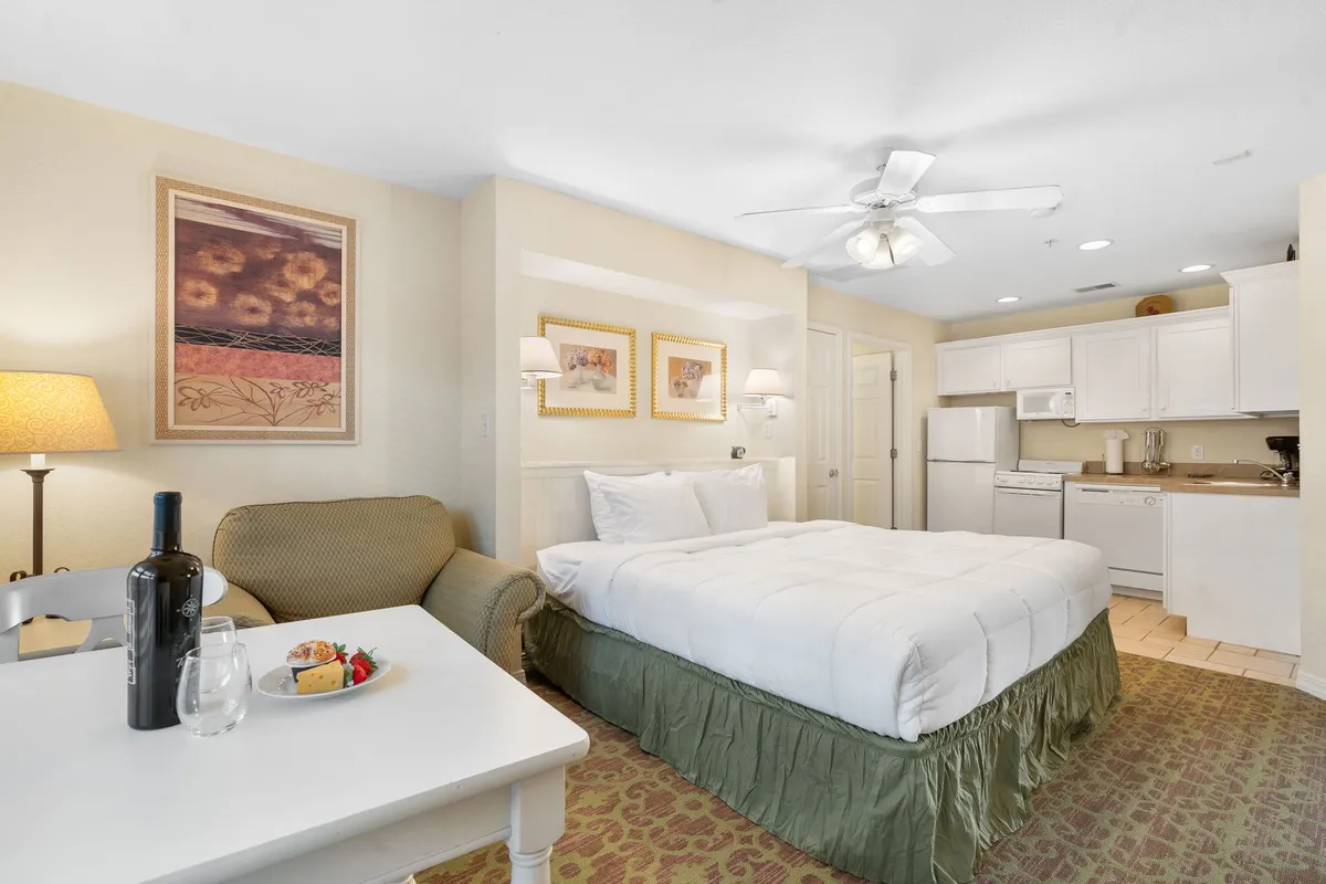 This inviting bedroom scene features a king-sized bed with crisp white linens and a green skirt, accompanied by a small dining table set with wine and fruit. Artwork on the walls and natural light from the window add to the comfortable, private ambiance.