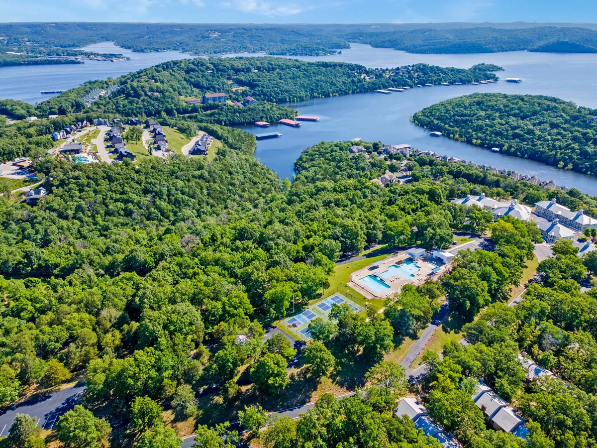A sweeping aerial view captures the resort's expansive grounds, with sparkling swimming pools, tennis courts, lush greenery, and the blue expanse of Table Rock Lake in the background. Residential buildings nestle between amenities, giving a sense of vibrant community life and direct access to outdoor fun.