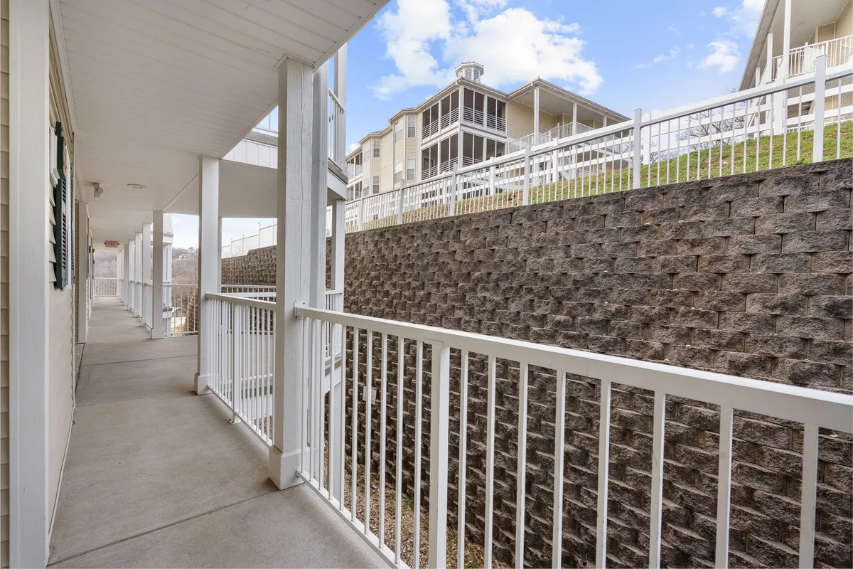 A calm, peaceful exterior walkway bordered by a stone wall and railing, with buildings and lush greenery in the background. The scene hints at walking paths and an inviting environment for leisurely strolls.