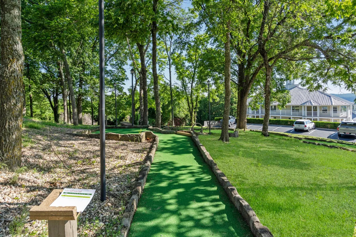 A charming mini-golf course winds through tall trees, with a signpost and picnic tables enhancing the playful, family-friendly atmosphere. The lush setting encourages outdoor gathering and lighthearted fun.