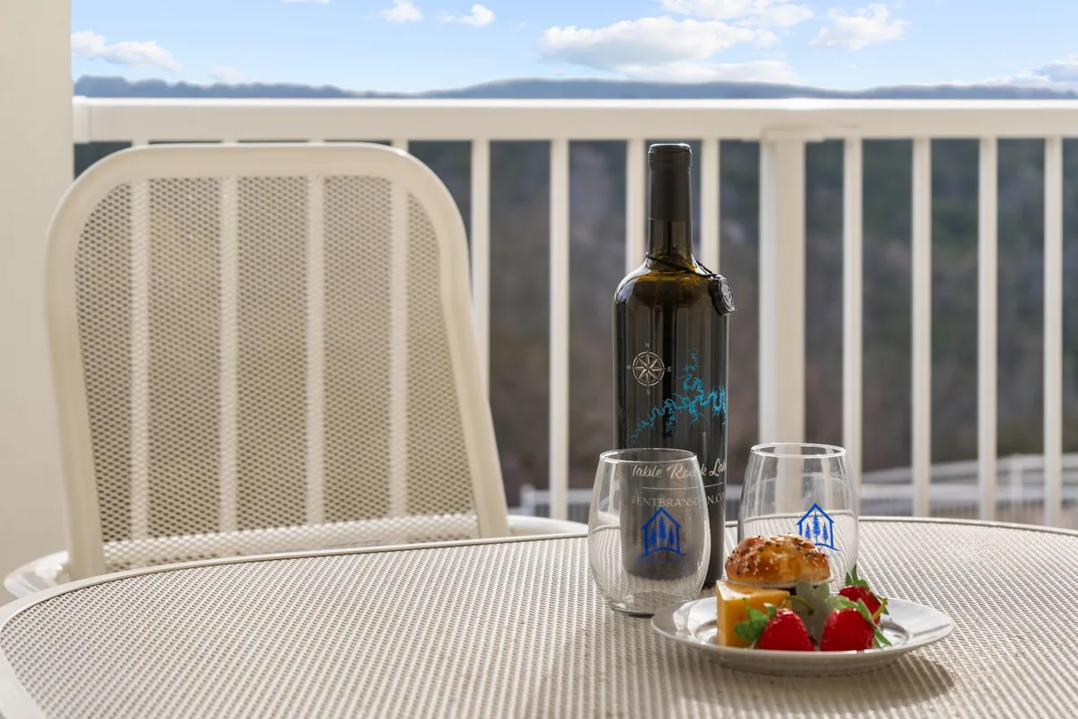 An exterior shot of a balcony or deck setup with a bottle of wine, two glasses, and a dessert plate. The setting is intimate and suggests a relaxing outdoor space with scenic views, emphasizing opportunities for guests to lounge outside.